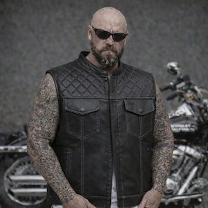 May include: A black leather vest with a diamond-stitched pattern on the shoulders and upper chest. The vest features two front pockets with button fastenings. The person in the image is wearing sunglasses and has tattoos on their arms. A motorcycle is in the background.
