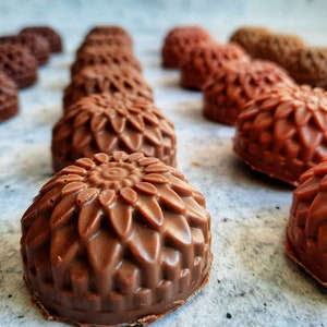May include: Close-up of several chocolate candies with a floral design. The candies are various shades of brown, from light to dark, and arranged in rows. The detailed design resembles a sunflower.