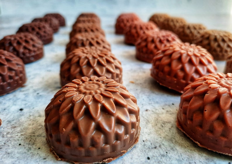 May include: Close-up of several chocolate candies with a floral design. The candies are various shades of brown, from light to dark, and arranged in rows. The detailed design resembles a sunflower.