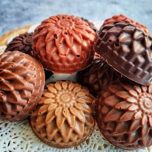 May include: An assortment of handmade soaps in various shades of brown, arranged on a white lace doily. The soaps are round with intricate floral designs, showcasing a range of chocolate and caramel hues. The soaps are a mix of light and dark shades.