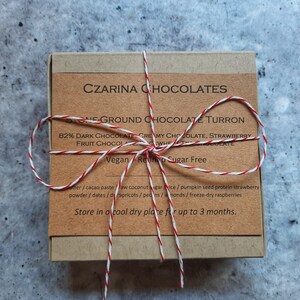 May include: A small, square box of Czarina Chocolates, tied with red and white twine. The label reads "Stone-Ground Chocolate Turron" and lists ingredients like dark chocolate and strawberries. The box is on a gray surface.