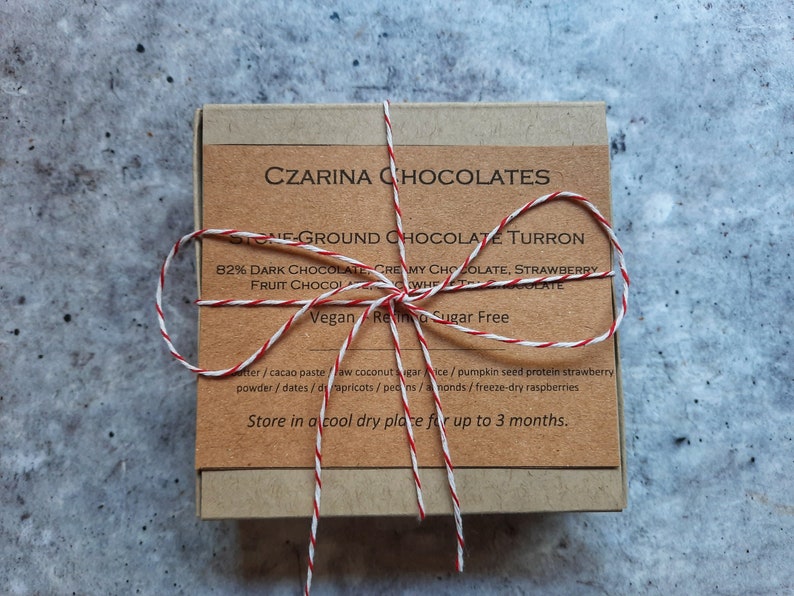 May include: A small, square box of Czarina Chocolates, tied with red and white twine. The label reads "Stone-Ground Chocolate Turron" and lists ingredients like dark chocolate and strawberries. The box is on a gray surface.
