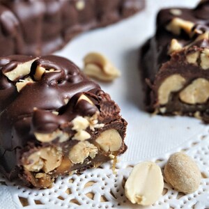 May include: Close-up of chocolate peanut butter bars with peanuts and caramel. The bars are on a white doily.
