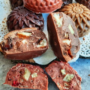 May include: Close-up of assorted chocolate candies with various shapes and fillings. Some are decorated with floral patterns, while others are filled with nuts and dried fruit. The candies are arranged on a wooden slice and a white doily.