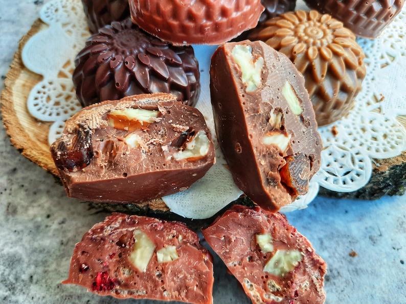 May include: Close-up of assorted chocolate candies with various shapes and fillings. Some are decorated with floral patterns, while others are filled with nuts and dried fruit. The candies are arranged on a wooden slice and a white doily.