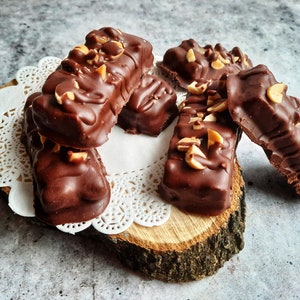 May include: A close-up of several chocolate peanut butter bars on a wooden slice with a white doily. The bars are covered in chocolate and have chopped peanuts on top.