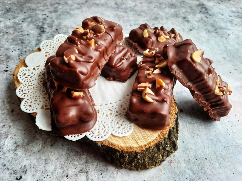 May include: A close-up of several chocolate peanut butter bars on a wooden slice with a white doily. The bars are covered in chocolate and have chopped peanuts on top.