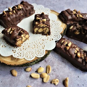 May include: A close-up of homemade chocolate peanut butter bars on a white doily and a wooden slice. The bars are covered in chocolate and topped with peanuts. Some peanuts are scattered on the table.