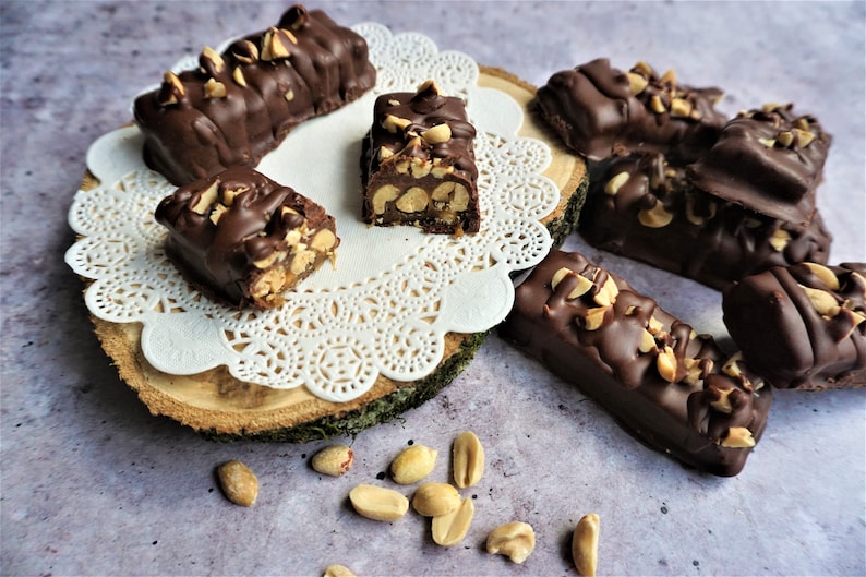 May include: A close-up of homemade chocolate peanut butter bars on a white doily and a wooden slice. The bars are covered in chocolate and topped with peanuts. Some peanuts are scattered on the table.