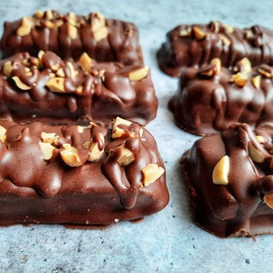 May include: Six chocolate peanut butter bars with chopped peanuts on top. The bars are arranged in two rows of three on a light blue surface.
