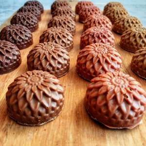 May include: Rows of handcrafted, flower-shaped soaps in varying shades of brown, from dark chocolate to light tan, are arranged on a wooden surface. The soaps have a detailed floral design and are likely made with natural ingredients.