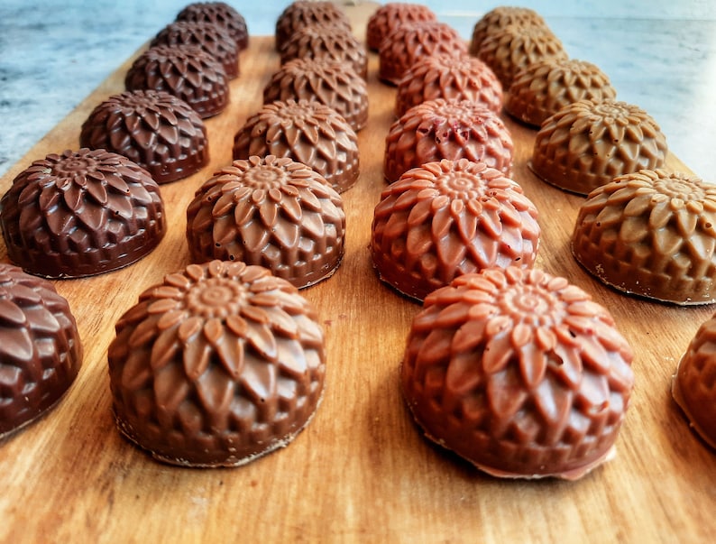May include: Rows of handcrafted, flower-shaped soaps in varying shades of brown, from dark chocolate to light tan, are arranged on a wooden surface. The soaps have a detailed floral design and are likely made with natural ingredients.
