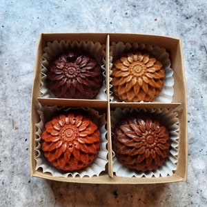 May include: A square cardboard box containing four chocolates, each molded in a flower shape. The chocolates vary in color from dark brown to light brown and are nestled in individual paper cups. The box sits on a mottled gray surface.
