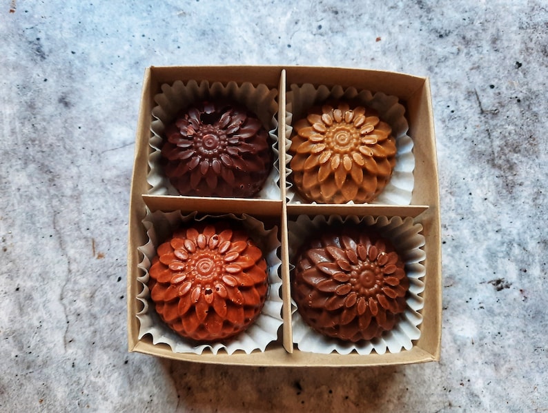 May include: A square cardboard box containing four chocolates, each molded in a flower shape. The chocolates vary in color from dark brown to light brown and are nestled in individual paper cups. The box sits on a mottled gray surface.