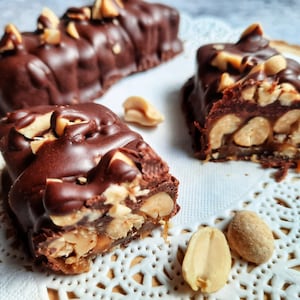 May include: Close-up of a chocolate peanut butter bar with peanuts and caramel. The bar is broken in half, revealing the layers of caramel and peanuts inside. The bar is sitting on a white doily.