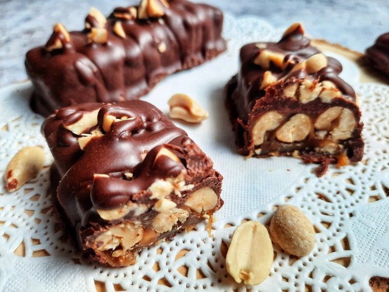 May include: Close-up of a chocolate peanut butter bar with peanuts and caramel. The bar is broken in half, revealing the layers of caramel and peanuts inside. The bar is sitting on a white doily.