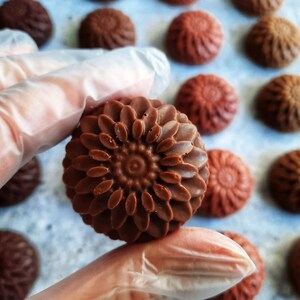 May include: Close-up of several chocolate candies with a floral design. The candies are various shades of brown, with one in the foreground held by a gloved hand. The candies have a detailed, textured surface, resembling a flower.