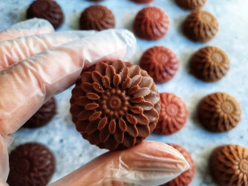 May include: Close-up of several chocolate candies with a floral design. The candies are various shades of brown, with one in the foreground held by a gloved hand. The candies have a detailed, textured surface, resembling a flower.