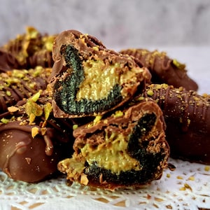May include: A close-up of chocolate-covered dates filled with a creamy filling. The dates are sprinkled with chopped pistachios and arranged on a white lace doily.