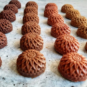 May include: An overhead shot displays rows of chocolate candies with a floral design. The candies vary in shades of brown, from dark to light, and are arranged on a light gray surface. The candies have a detailed, textured appearance.