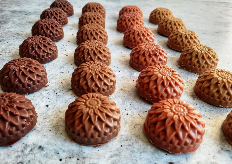 May include: An overhead shot displays rows of chocolate candies with a floral design. The candies vary in shades of brown, from dark to light, and are arranged on a light gray surface. The candies have a detailed, textured appearance.