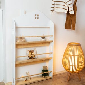 Könnte beinhalten: Ein weißes, hausförmiges Bücherregal mit Holzregalen und Kinderartikeln. Ein Holzspielzeugzug, ein Steinstapel, ein Buch, ein Teddybär und ein Stoffhase sind ausgestellt. Ein weißer Pullover und eine braune Latzhose hängen an der Wand.