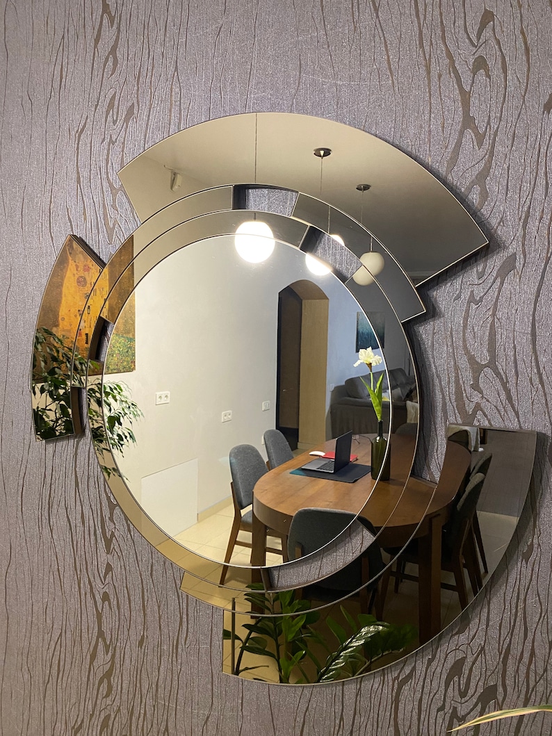 K&ouml;nnte beinhalten: Ein gro&szlig;er, runder, moderner Spiegel mit geometrischem Design. Der Spiegel besteht aus mehreren Glasschichten, die ein einzigartiges und stilvolles Aussehen erzeugen. Der Spiegel spiegelt ein Esszimmer mit einem Holztisch und St&uuml;hlen wider.