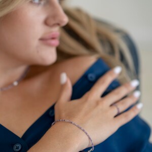 May include: A woman wearing a dark blue shirt with a delicate purple beaded bracelet on her wrist. She is wearing a silver necklace with a small pearl pendant.