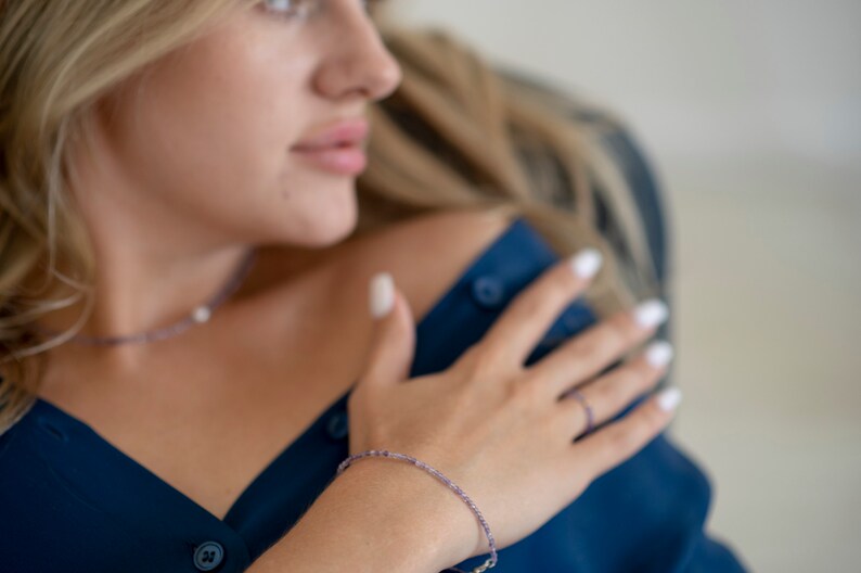 May include: A woman wearing a dark blue shirt with a delicate purple beaded bracelet on her wrist. She is wearing a silver necklace with a small pearl pendant.