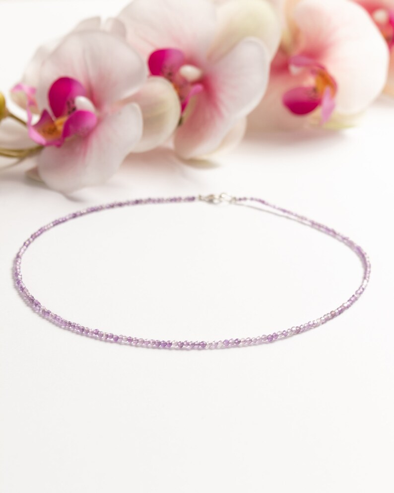 May include: A delicate necklace made with small, light purple amethyst beads. The necklace is on a white background with pink orchid flowers.