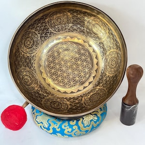 May include: A golden singing bowl with a flower of life design and a wooden striker. The bowl is resting on a blue and gold cushion.