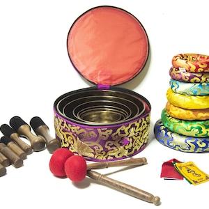 May include: A set of seven singing bowls with a purple and gold embroidered carrying case, two wooden mallets with red felt heads, and seven colorful cushions. The bowls are nested inside each other and the cushions are stacked in a rainbow pattern.