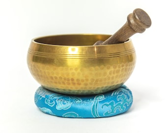 May include: A gold singing bowl with a wooden striker, resting on a blue and white patterned cushion.