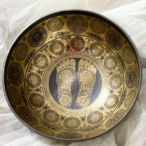May include: A large, round singing bowl with a black interior and a gold exterior. The bowl is decorated with intricate patterns and designs, including two stylised footprints in the centre.