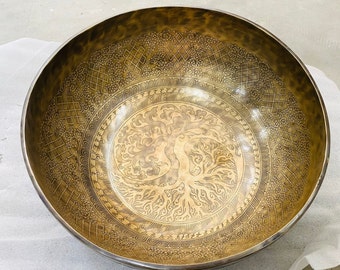 May include: A large, round, brass singing bowl with intricate etched designs. The bowl features a central tree of life motif with swirling branches and roots.