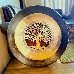 Puede incluir: Un gong grande y redondo con un borde exterior oscuro y un centro dorado. El centro presenta un diseño detallado de árbol de la vida. El gong está hecho de metal y probablemente se usa para la curación por sonido o la meditación.