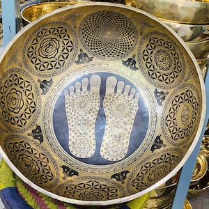 May include: A large, golden singing bowl with intricate designs. The bowl features a black centre with two stylised footprints, surrounded by a mandala pattern. The bowl is decorated with a variety of geometric shapes and symbols.