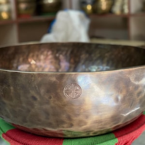 May include: A large, bronze-colored singing bowl with a hammered texture. The bowl has a circular design etched into the side. The bowl is resting on a red and green cushion. The background is blurred.