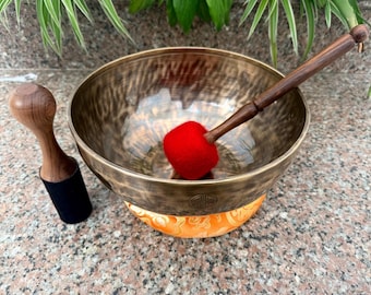 May include: A bronze-colored metal singing bowl with a hammered texture and a circular emblem on the side. It includes a wooden mallet with a bright red felt head resting inside, and a separate wooden mallet with a black felt base beside it.