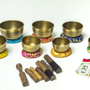 May include: Set of seven golden singing bowls with intricate designs, each resting on a colourful cushion. The bowls come with seven wooden strikers and a set of prayer flags.