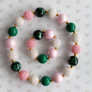 May include: A necklace and bracelet set with pink, white, green, and gold beads. The necklace and bracelet have a variety of bead styles, including smooth, glittery, and textured beads. The beads are strung together with gold-coloured connectors.