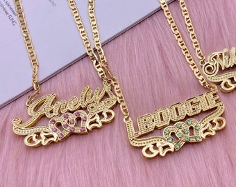 May include: Three gold necklaces with nameplates featuring the names "Anelly", "LBODGIE", and "Nikkibanks" with jeweled hearts. The necklaces are on a pink fuzzy surface.