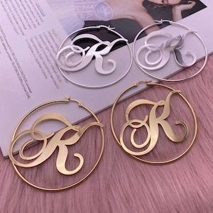 May include: Two pairs of hoop earrings with a large, cursive letter "R" cut out of the metal. One pair is gold and the other is silver.