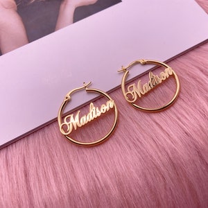 May include: A pair of gold hoop earrings with the name "Madison" written in cursive on each earring.