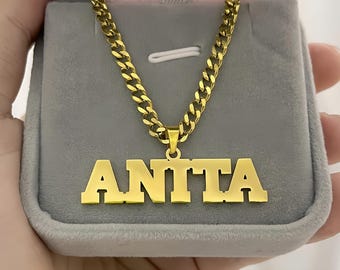 Puede incluir: Un collar dorado con el nombre "ANITA" en letras negritas y mayúsculas. El collar tiene una gruesa cadena de eslabones y se muestra en una caja de joyería gris abierta. También se ve un paño de limpieza rosa.