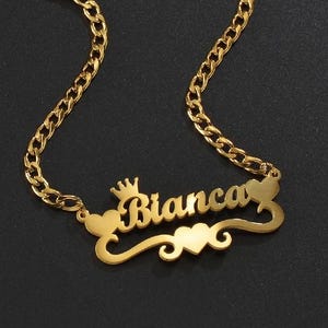 Può includere: Una collana color oro con il nome "Bianca" in corsivo. La collana presenta una corona sopra il nome e dettagli a forma di cuore. La catena è a maglia.