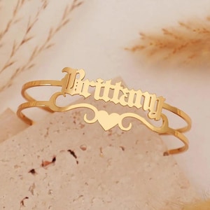May include: Gold-colored bracelet with the name "Brittany" in an elegant font. The bracelet features a heart-shaped design and decorative swirls. The bracelet is a stylish accessory.