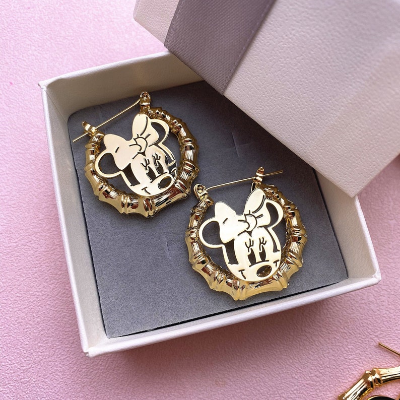 May include: A pair of gold hoop earrings featuring a cutout design of a cartoon character with a bow. The earrings are presented in a white box with a grey ribbon.