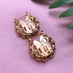 May include: A pair of gold-toned hoop earrings featuring a cut-out design of Minnie Mouse's face. The earrings are made from a bamboo-like material.