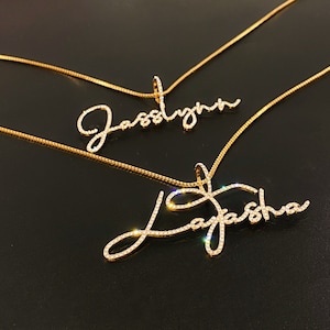 May include: Two gold-colored name necklaces. One necklace spells out "Jasslynn" and the other spells out "Layasha". The names are written in a cursive font and are encrusted with small, sparkling crystals. The necklaces are on a dark background.
